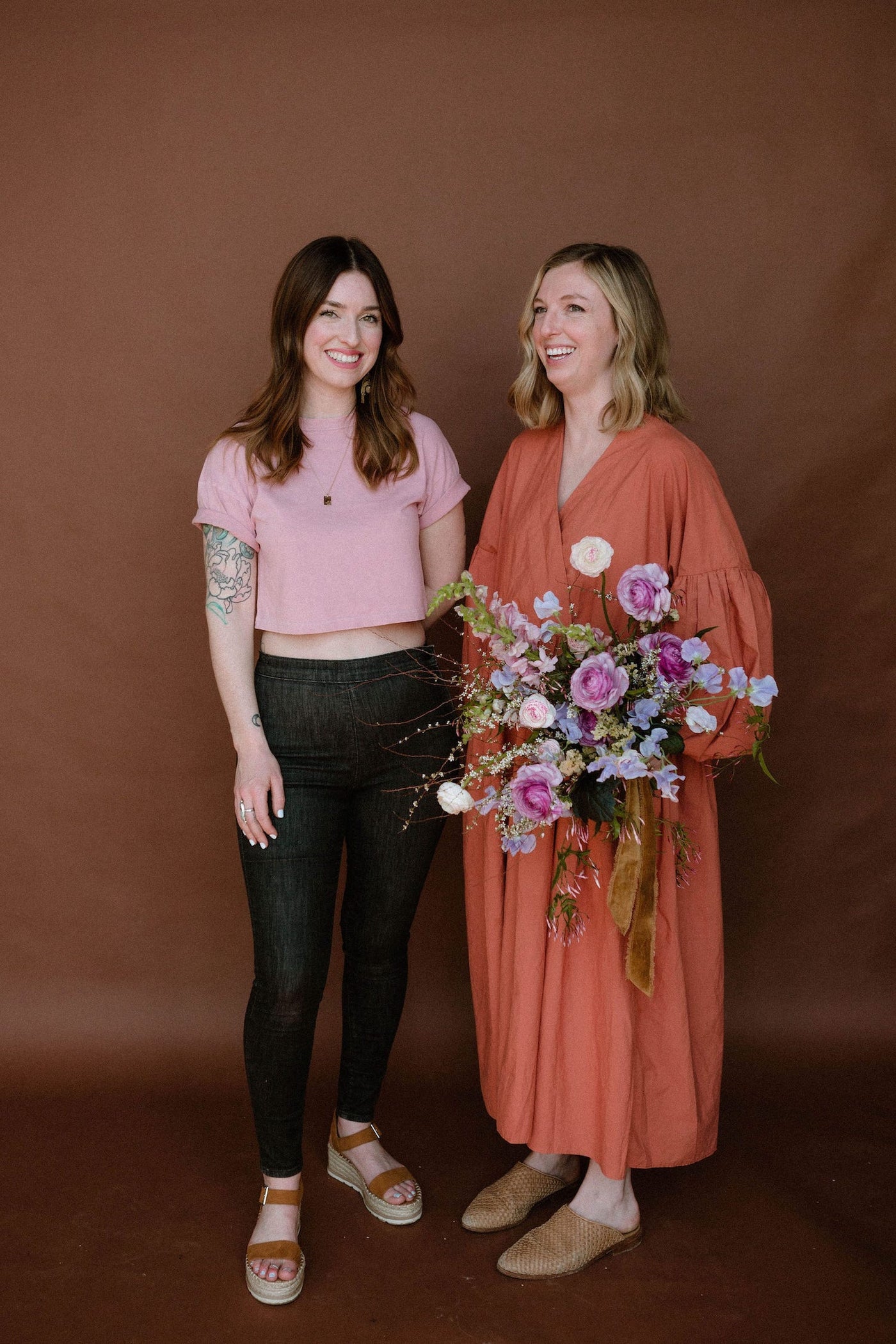 Katie and Jenny of Anthousai. Katie is holding a bouquet and they are standing behind a solid background.